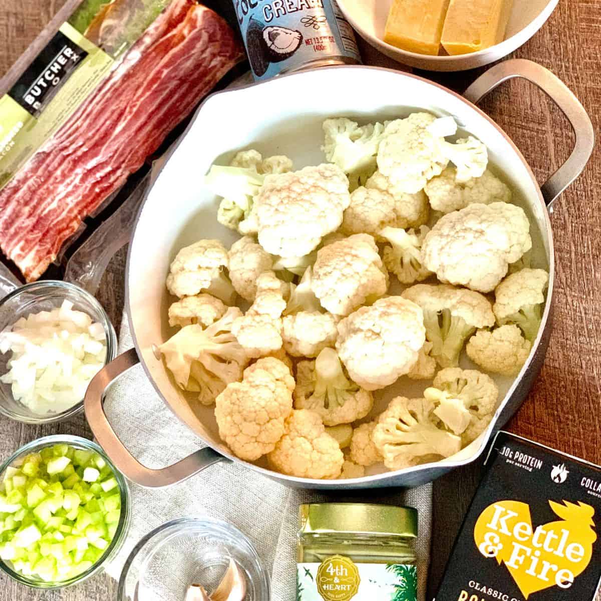 Ingredients displayed together to make the dairy free cauliflower soup, including chopped cauliflower florets in a large pan with chicken bone broth, ghee, garlic cloves, celery, onions, bacon strips and coconut cream.