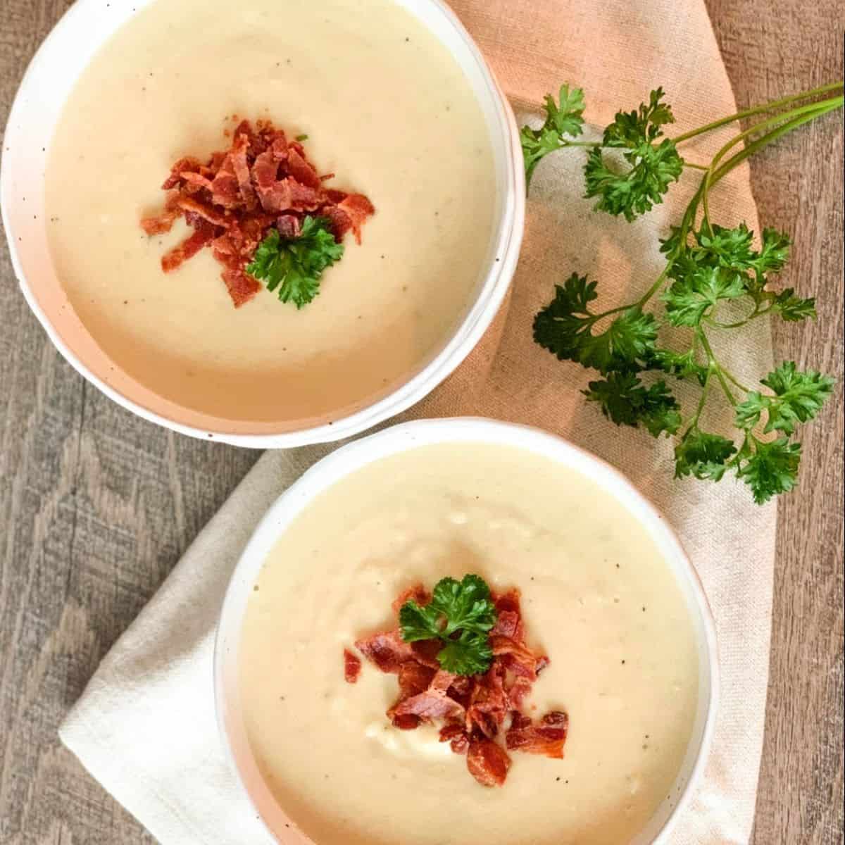 Two white bowls filled with dairy free cauliflower soup with a garnish of crispy bacon and fresh parsley.