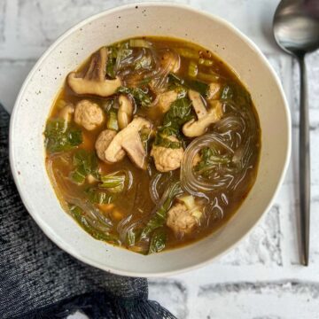 White speckled bowl filled with glass noodle ramen with ground pork meatballs, shiitake mushrooms, baby bok choy and green onions in a lemongrass ginger beef bone broth. There's a dark napkin in the bottom left corner and a big gray spoon in the top right corner.