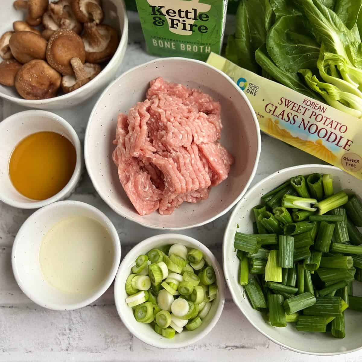 All ingredients for the glass noodle ramen soup laid out in white speckled bowls. this includes the ground pork, sweet potato glass noodles, green parts of the scallions and white parts sliced differently, baby bok choy, mushrooms, sesame oil, rice vinegar and bone broth.