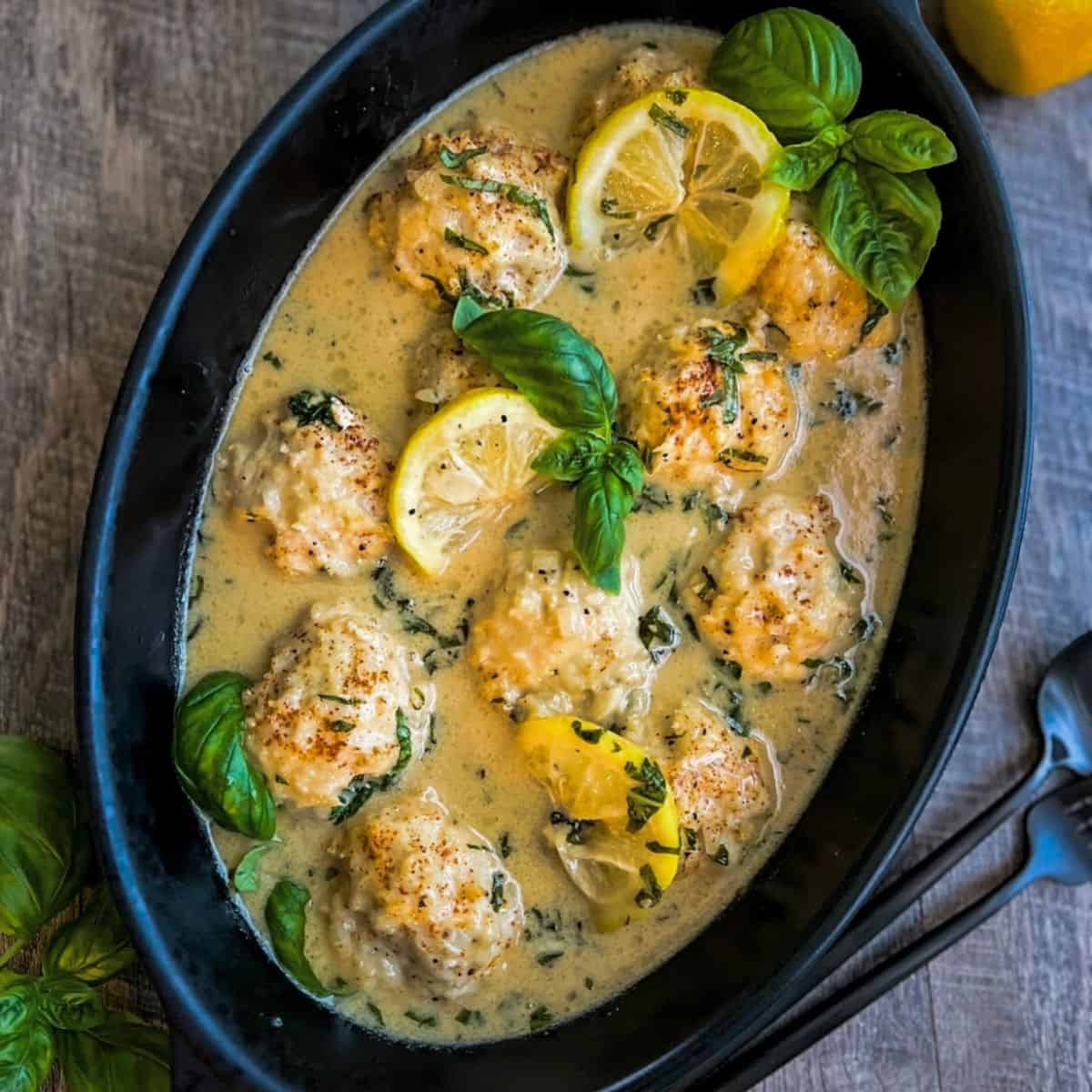 Black serving platter with gluten free chicken meatballs laid out in a sauce with lemon slices and fresh sprigs of basil.