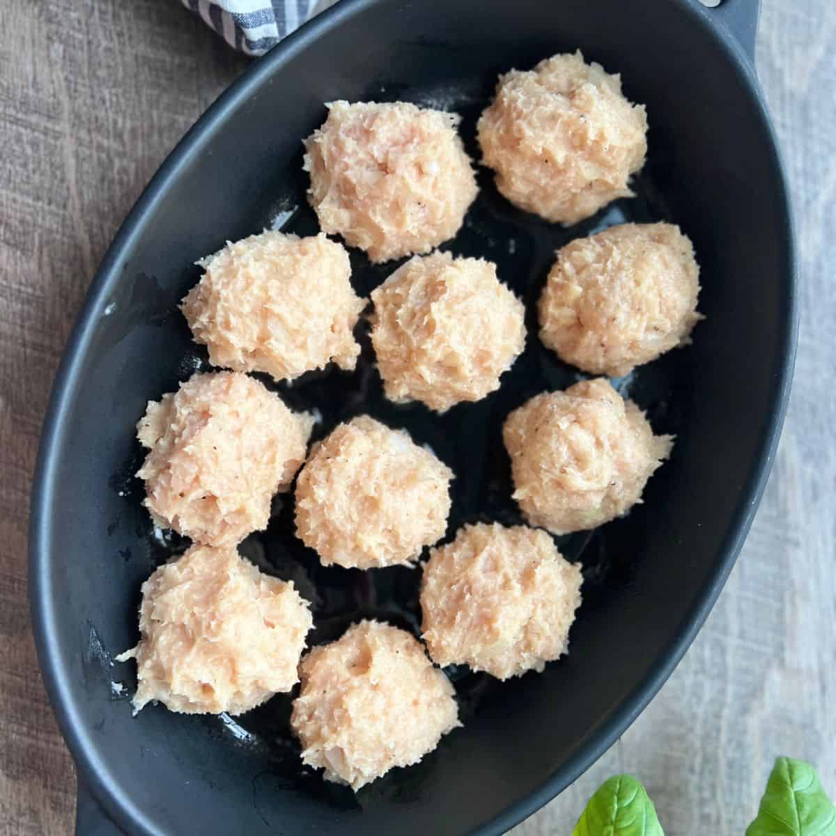 Rolled out ground chicken meatballs in a black dish before going into the oven with a few basil leaves peaking out on the bottom.