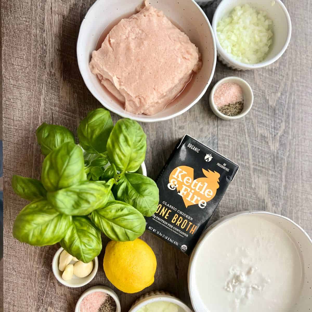 Ingredients laid out on the countertop for gluten free chicken meatballs including Kettle & Fire bone broth, fresh basil leaves, ground chicken, diced onion, salt and pepper, a whole lemon, garlic cloves and coconut cream in white bowls.