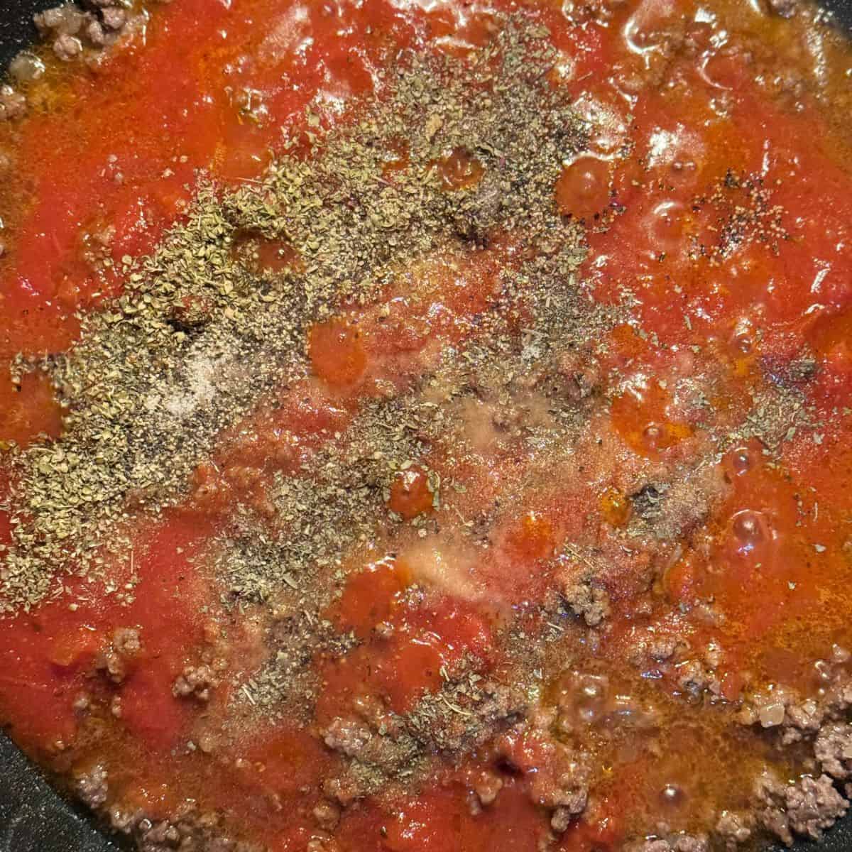 Bolognese sauce cooking in a large pan with the tomato basil sauce and spices sprinkled on top being ready to mix altogether.