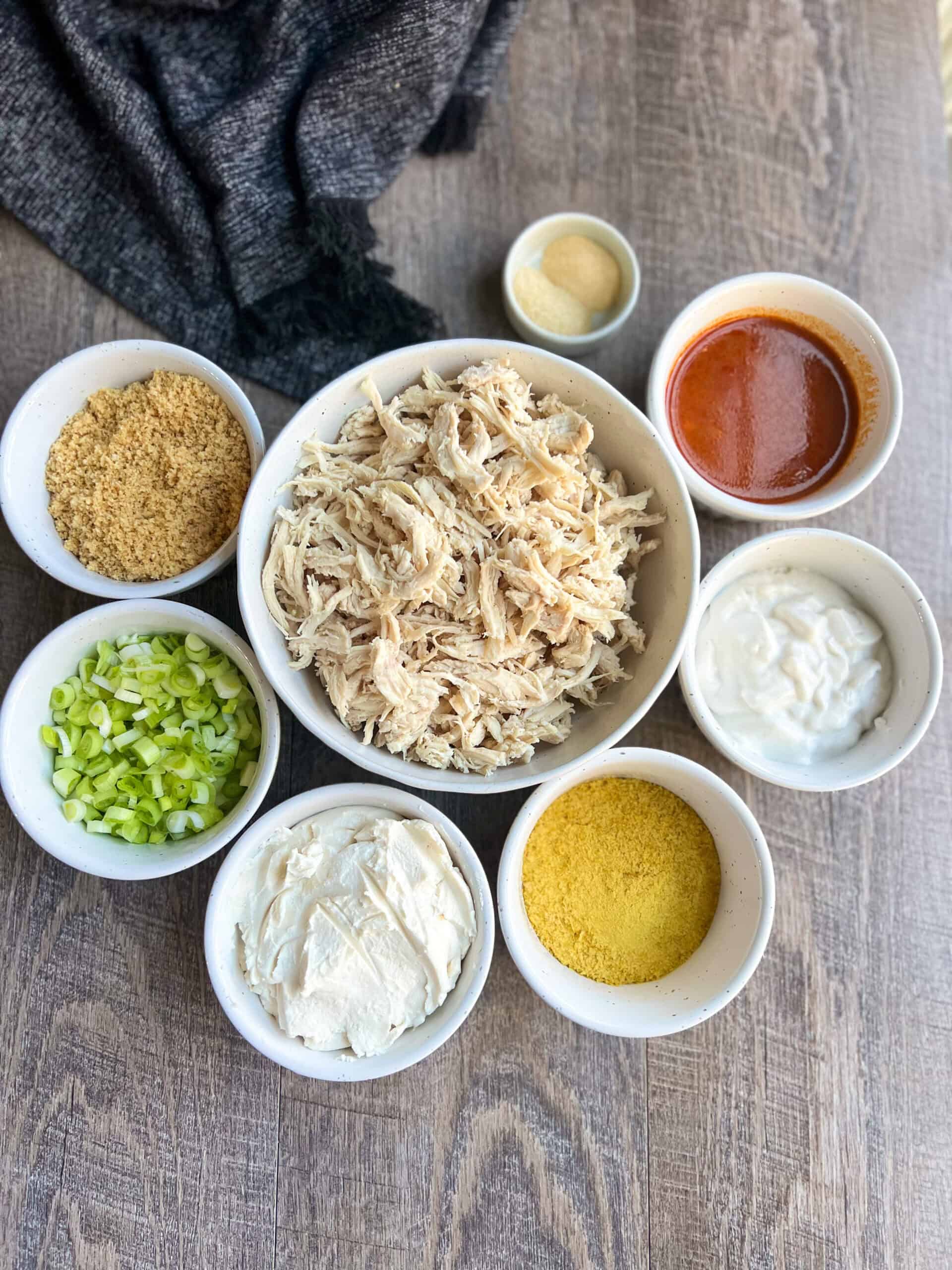 Ingredients for the paleo buffalo chicken dip are laid out in white bowls with the shredded chicken in the largest bowl in the center, surrounded by garlic and onion powder, buffalo sauce, dairy free sour cream, nutritional yeast, dairy free cream cheese, chopped scallions and pork panko.