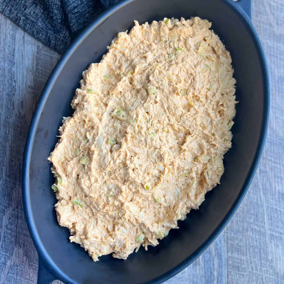 The base of the buffalo chicken dip is spread evenly in a black oven-safe baking dish and is ready to go into the oven.