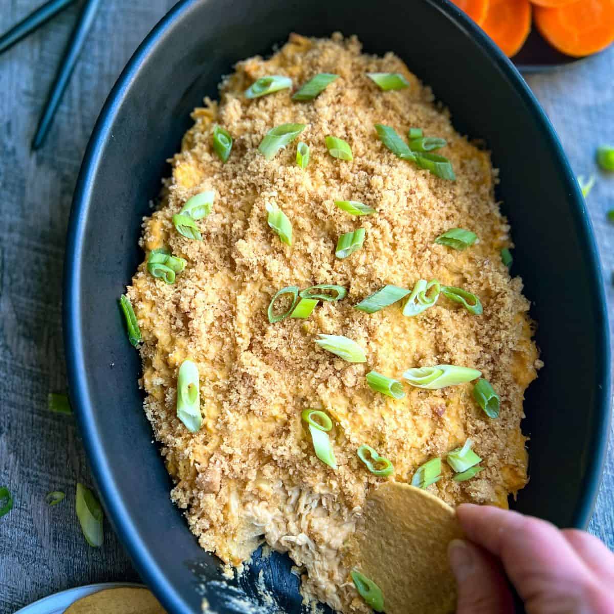 A black oven-safe baking dish with buffalo chicken dip spread evenly garnished with scallions and pork panko ready to be enjoyed. There is a hand with a tortilla chip dipping into the dip at the bottom to enjoy a bite.