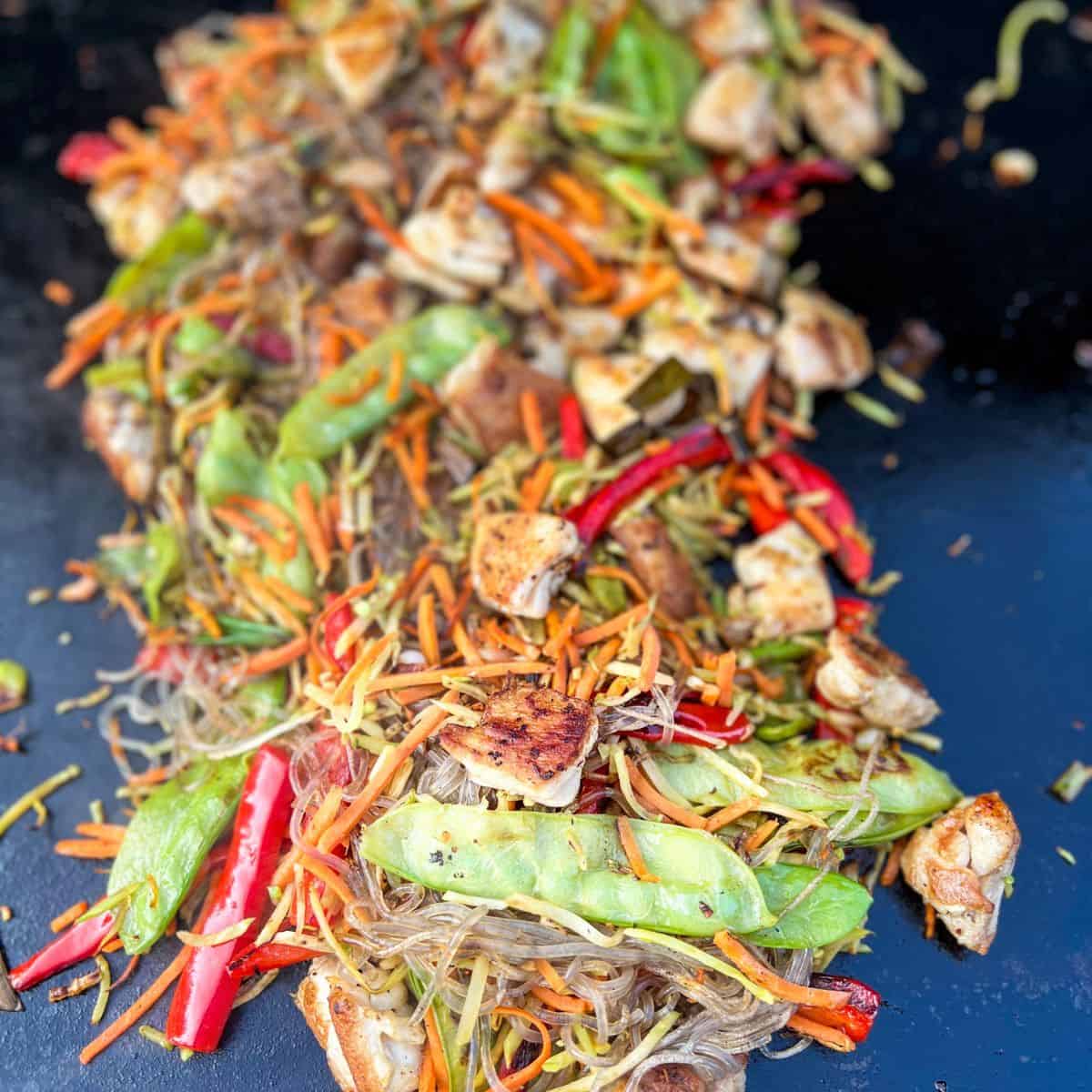 Chopped vegetables and chicken all mixed together on a blackstone grill top mixed with glass noodles.