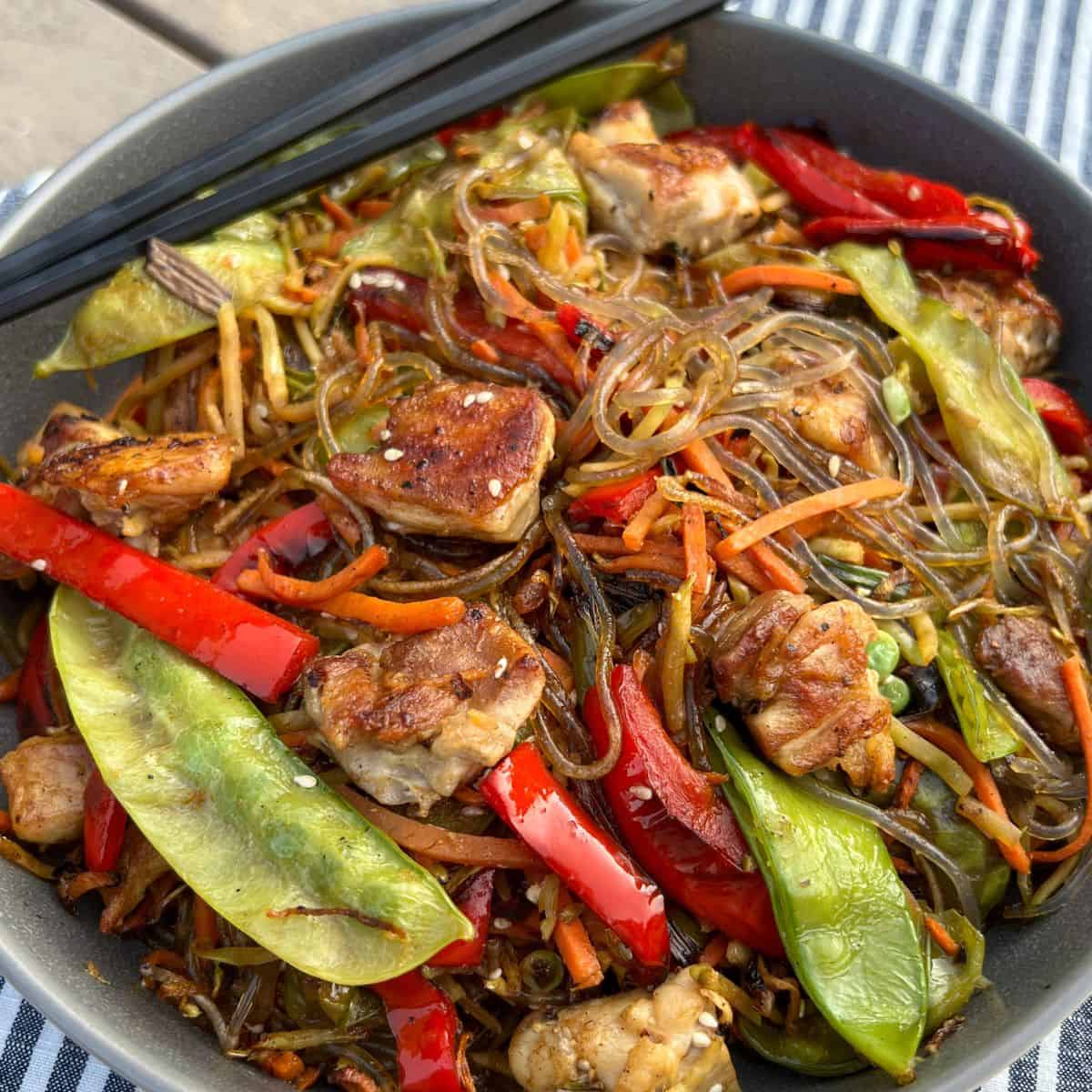 Presentation of Stir Fried Glass Noodles with colorful grilled vegetables and chicken in a grey bowl with chopsticks.