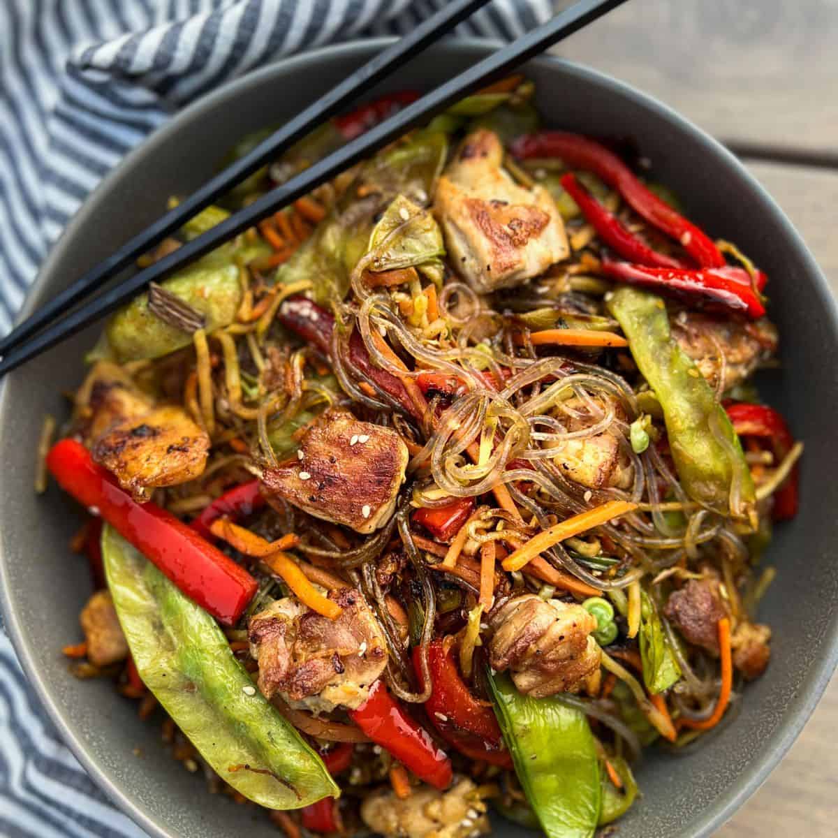 Presentation of Stir Fried Glass Noodles with colorful grilled vegetables and chicken in a grey bowl with chopsticks.